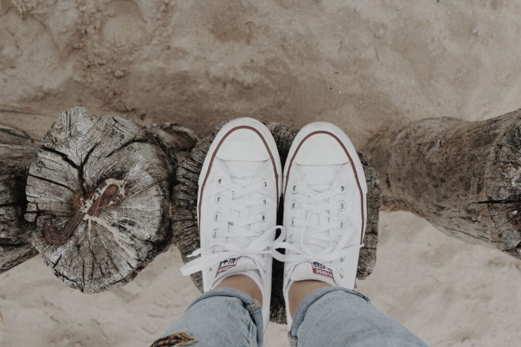 Meitenes pēdas baltajos converse apavos uz koka staba virsmas pludmalē