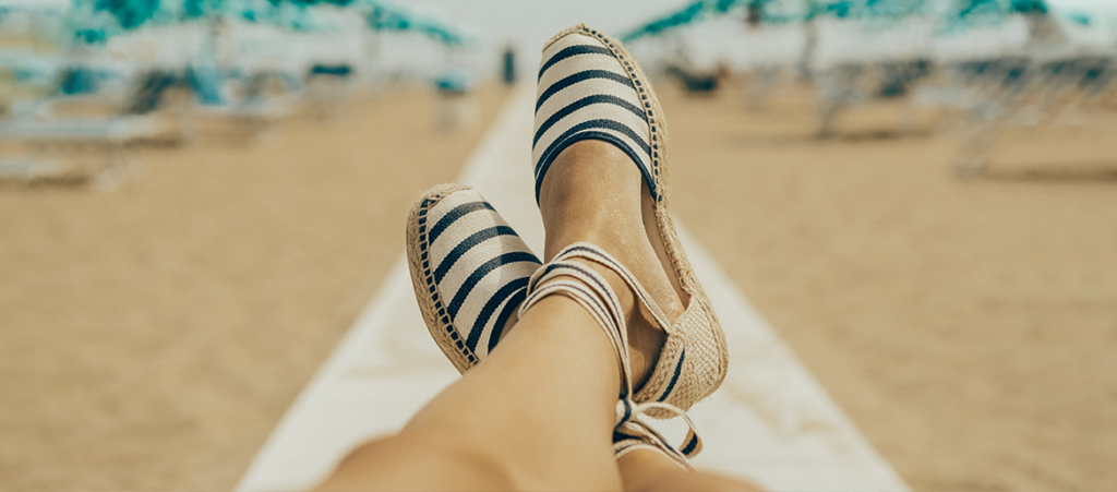 Sakrustotas sievietes kājas pludmalē, kurās ir sasienamas ap potīti sandales ar baltām un tumši zilām svītrām