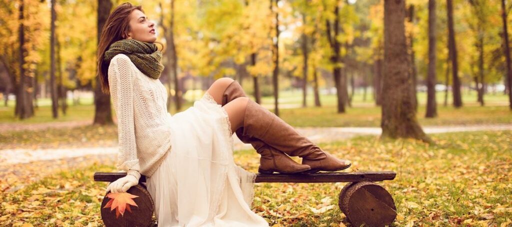 Sieviete sēž uz parka soliņa baltā kleitā un gaišos, garos zābakos ar platu stulma augšdaļu.
