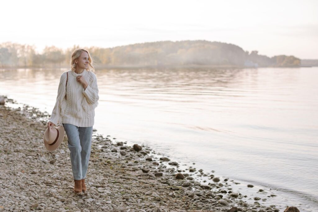 Sieviete baltā džemperī un kamieļkrāsas kovboju apavos pastaigājas gar ezera krastu.