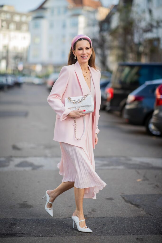 Sieviete tērpusies gaiši rozā krāsas kleitā ar lielizmēra žaketi tādā pašā krāsā un baltiem aksesuāriem, kā rokassomiņa un augstpapēžu sandales