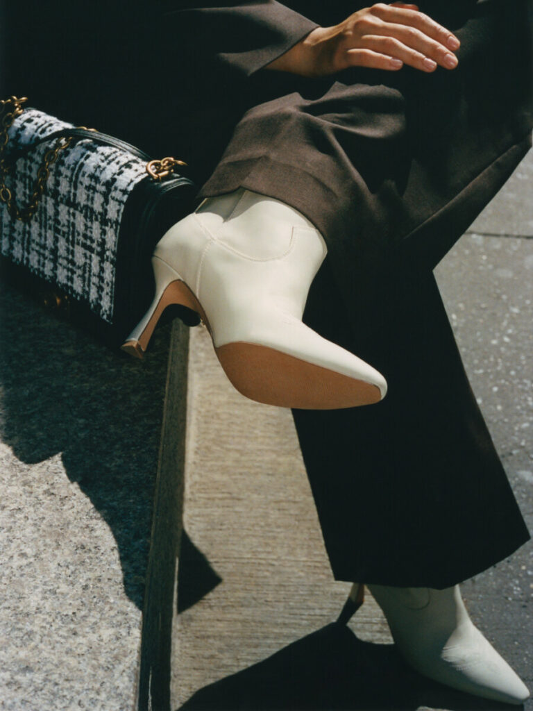 Modele ar Nine West zīmola melnbaltu somiņu ar rūtiņu rakstu un baltiem īsiem zābakiem un tievu papēdi