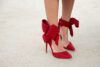 Sarkanas augstpapēžu kurpes ar smailu purngalu un bantes sasējumu potītes daļā, kas atgādina svētku dāvanas noskaņās