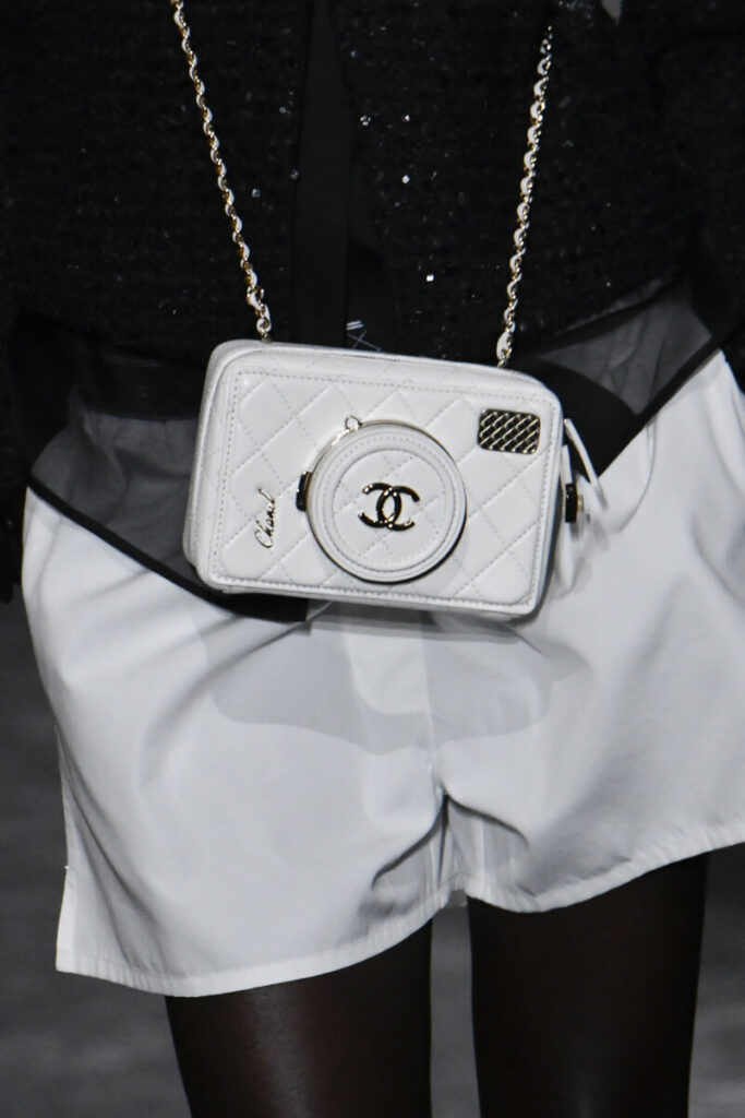 Modele melnbaltajā tērpā ar neliela izmēra baltas krāsas Chanel zīmola rokassomu ar ķēdīti